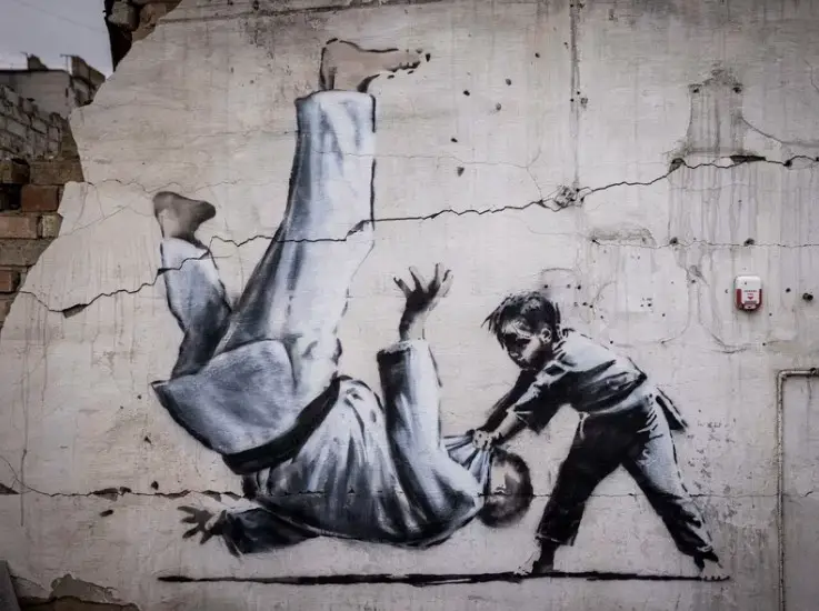 Graffiti of a child throwing a man on the floor in judo clothing is seen on a wall amid damaged buildings